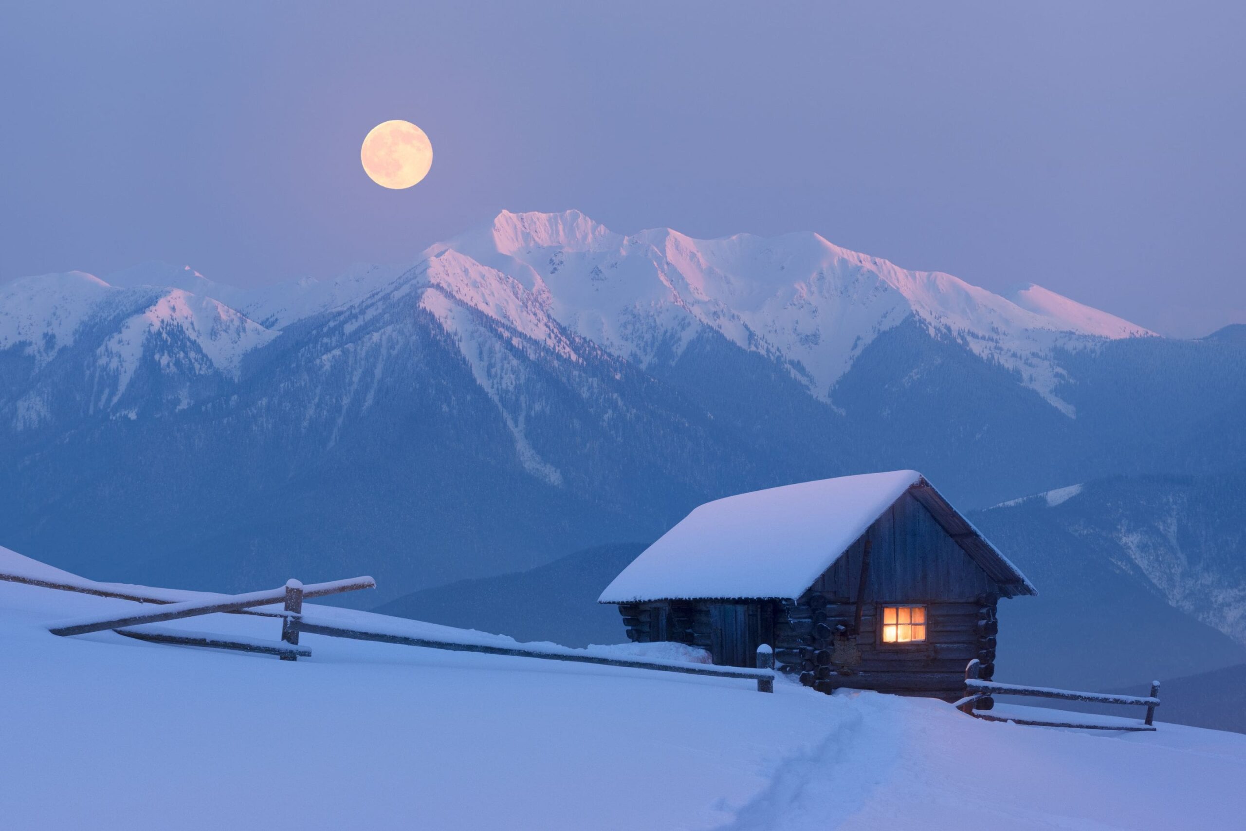 small cabin covered in snow with snowy mountains and a full moon in the background
