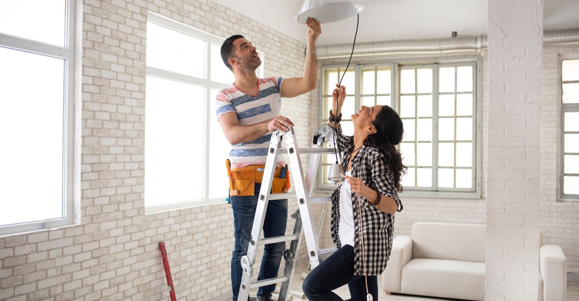 A man and woman stand on a ladder, each holding a light bulb, ready to replace the old one above them