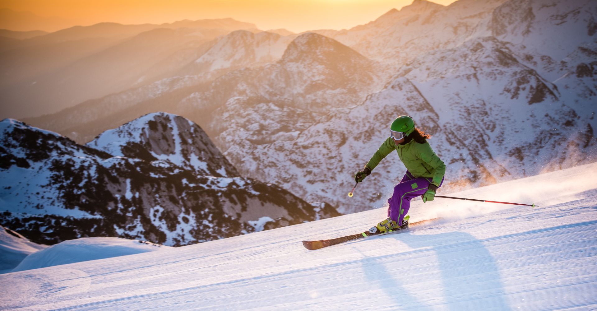 A person skis down a snowy mountain as the sun sets, illuminating the sky with warm hues of orange and pink