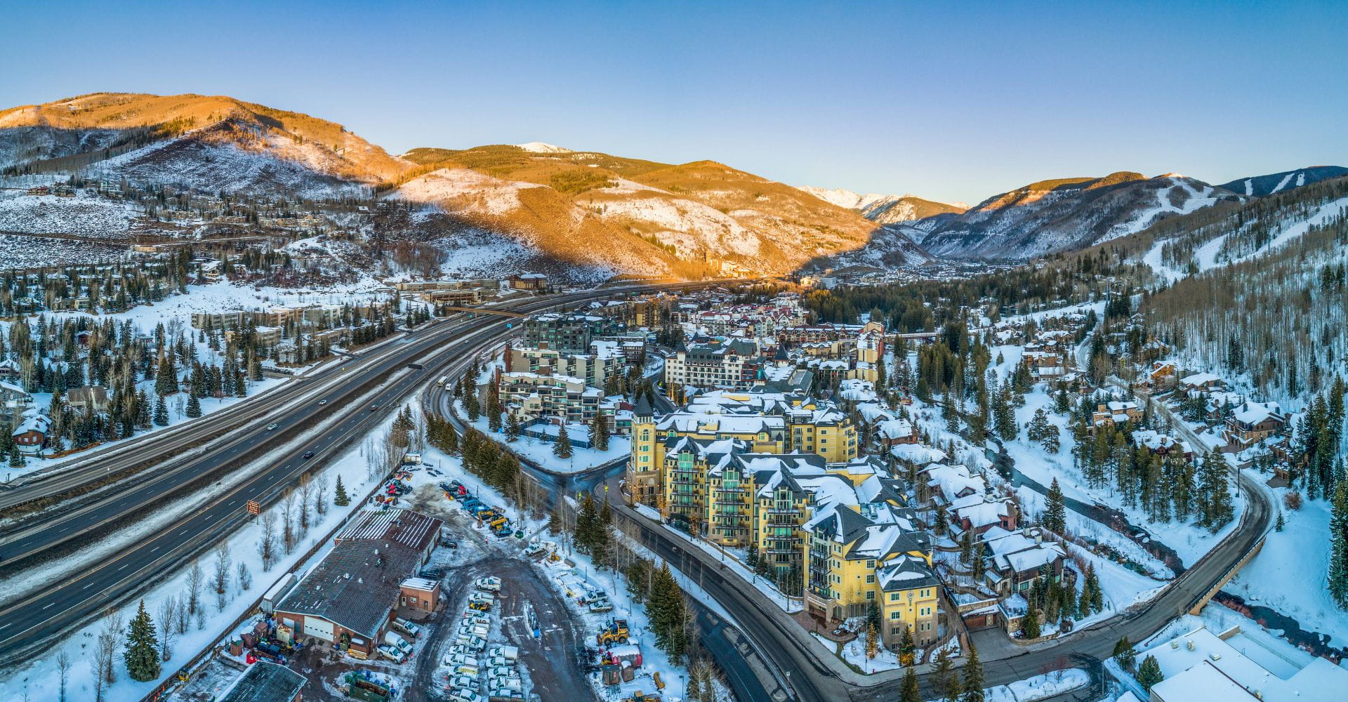 Aerial view of a snowy mountain town at sunrise. Buildings and highways are nestled between forested hills. The scene is serene and picturesque.